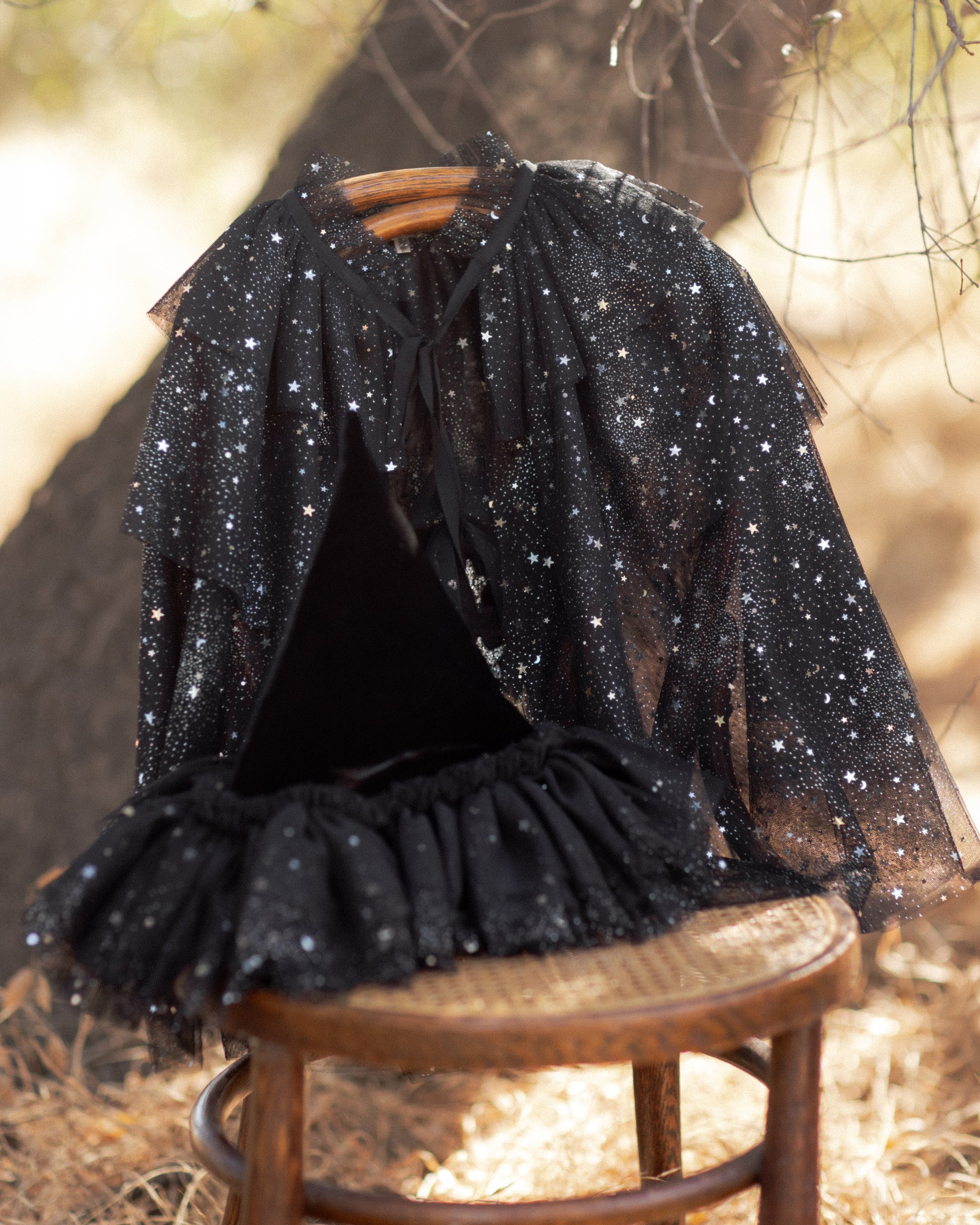A Noralee Tulle Cape in black, adorned with silver stars, hangs on a chair outdoors—ideal for dress-up or special occasions. A coordinating black pointed hat and skirt are also displayed on the chair.