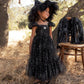 A young girl wearing Noralee’s Girls’ Clara Dress in Silver Stars—a black tulle dress adorned with silver star patterns and a smocked bodice—along with a large, matching hat, stands outdoors on dry grass beside a stool draped with coordinating pieces.