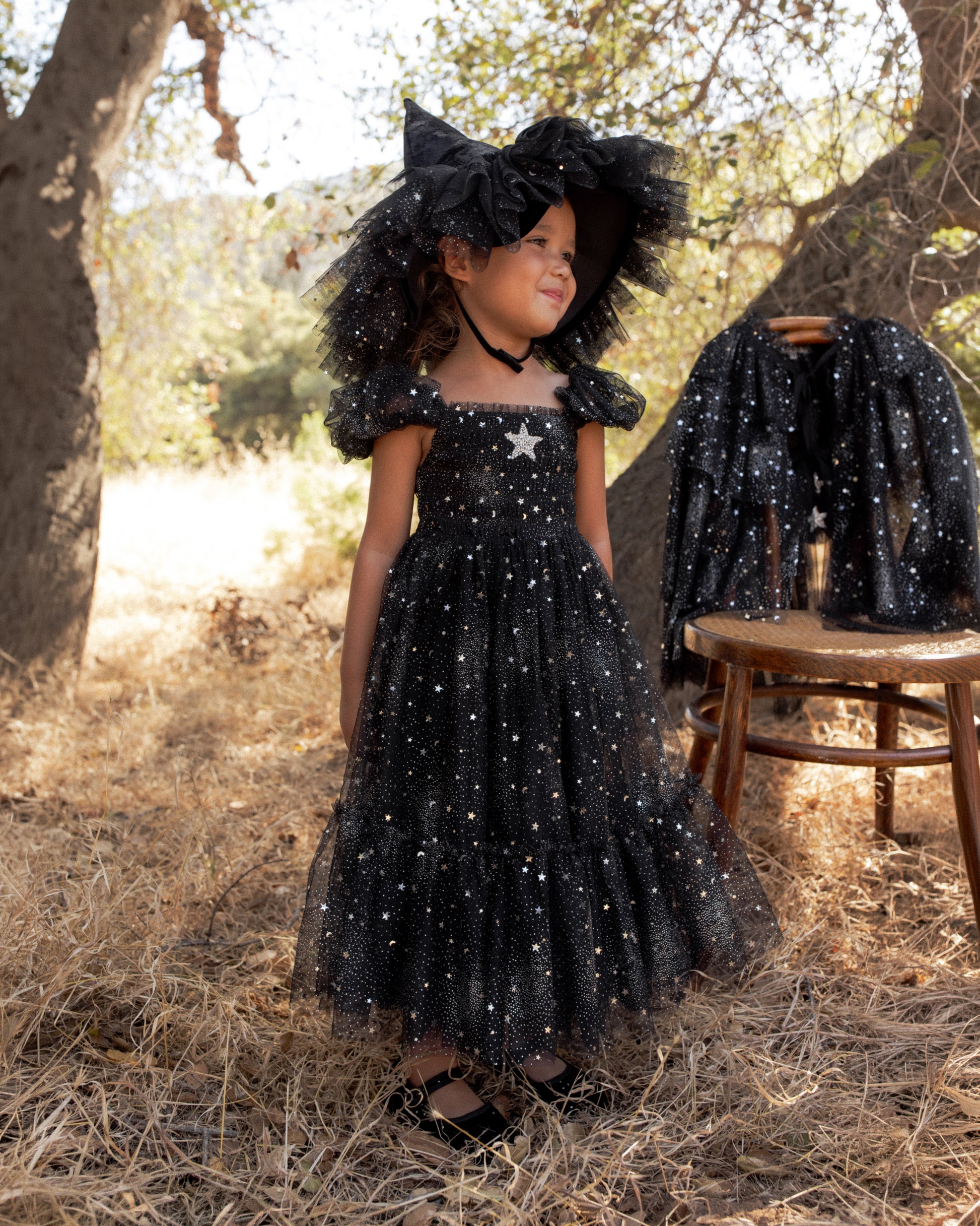 A young girl wearing Noralee’s Girls’ Clara Dress in Silver Stars—a black tulle dress adorned with silver star patterns and a smocked bodice—along with a large, matching hat, stands outdoors on dry grass beside a stool draped with coordinating pieces.