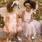 Two young girls, dressed in Noralee Wings fairy costumes, sit on grass covered in autumn leaves, smiling at each other and enjoying imaginative play.