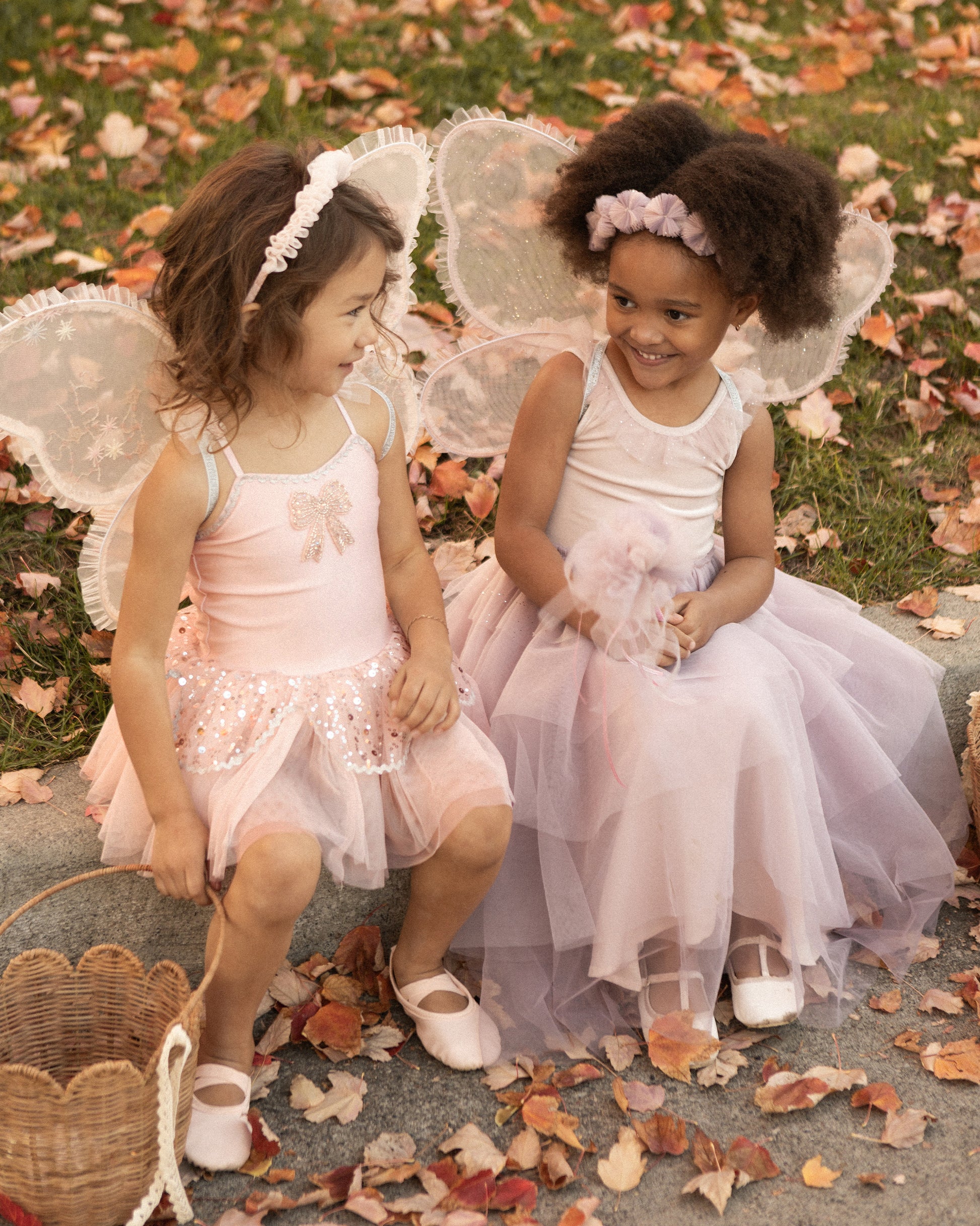 Two young girls, dressed in Noralee Wings fairy costumes, sit on grass covered in autumn leaves, smiling at each other and enjoying imaginative play.
