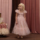 A young child wearing the Noralee Girls’ Clara Dress in light pink with a floral pattern and smocked bodice, paired with a tiara, stands on a wooden floor in front of pink curtains, looking to the side.