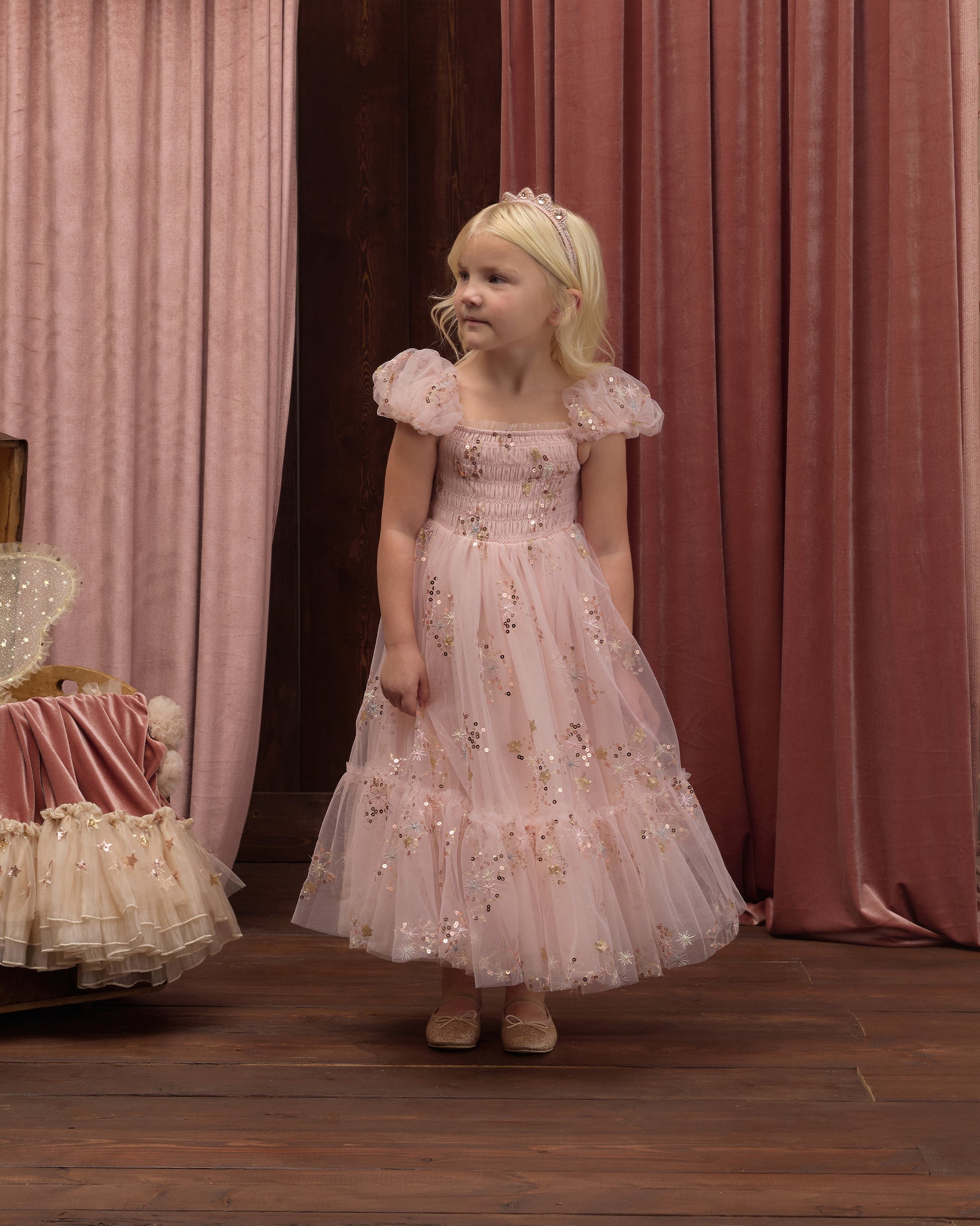 A young child wearing the Noralee Girls’ Clara Dress in light pink with a floral pattern and smocked bodice, paired with a tiara, stands on a wooden floor in front of pink curtains, looking to the side.