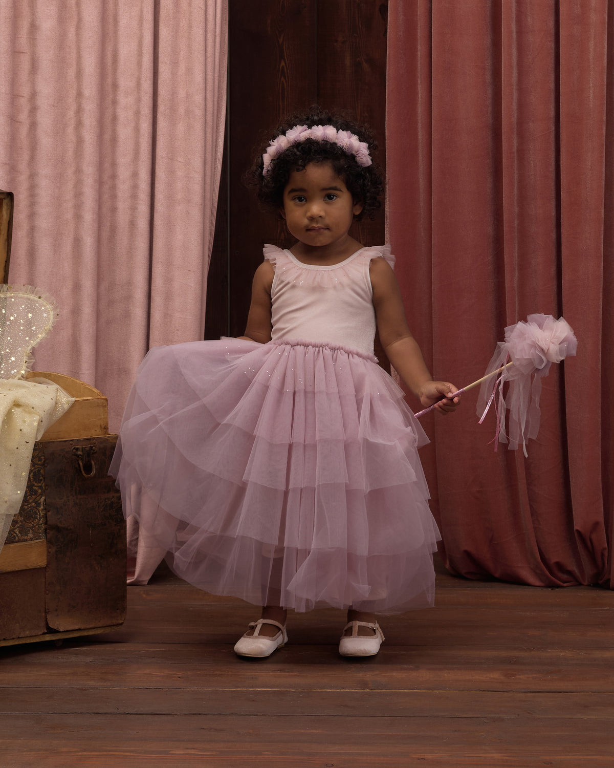 A young girl wears the Noralee Girls’ Pixie Dress, featuring a velvet bodice and tulle skirt, as she stands on a wooden floor holding a matching wand, with pink curtains in the background.