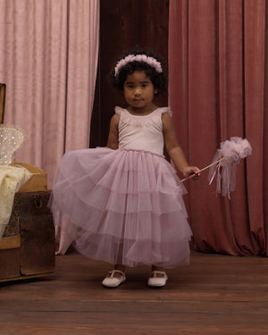 A young girl wears the Noralee Girls’ Pixie Dress, featuring a velvet bodice and tulle skirt, as she stands on a wooden floor holding a matching wand, with pink curtains in the background.