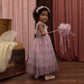 A young girl in a pink dress and headband holds the Noralee Pom Pom Crown + Wand, standing on a wooden floor with pink curtains and an open trunk in the background, ready for a magical dress-up adventure.