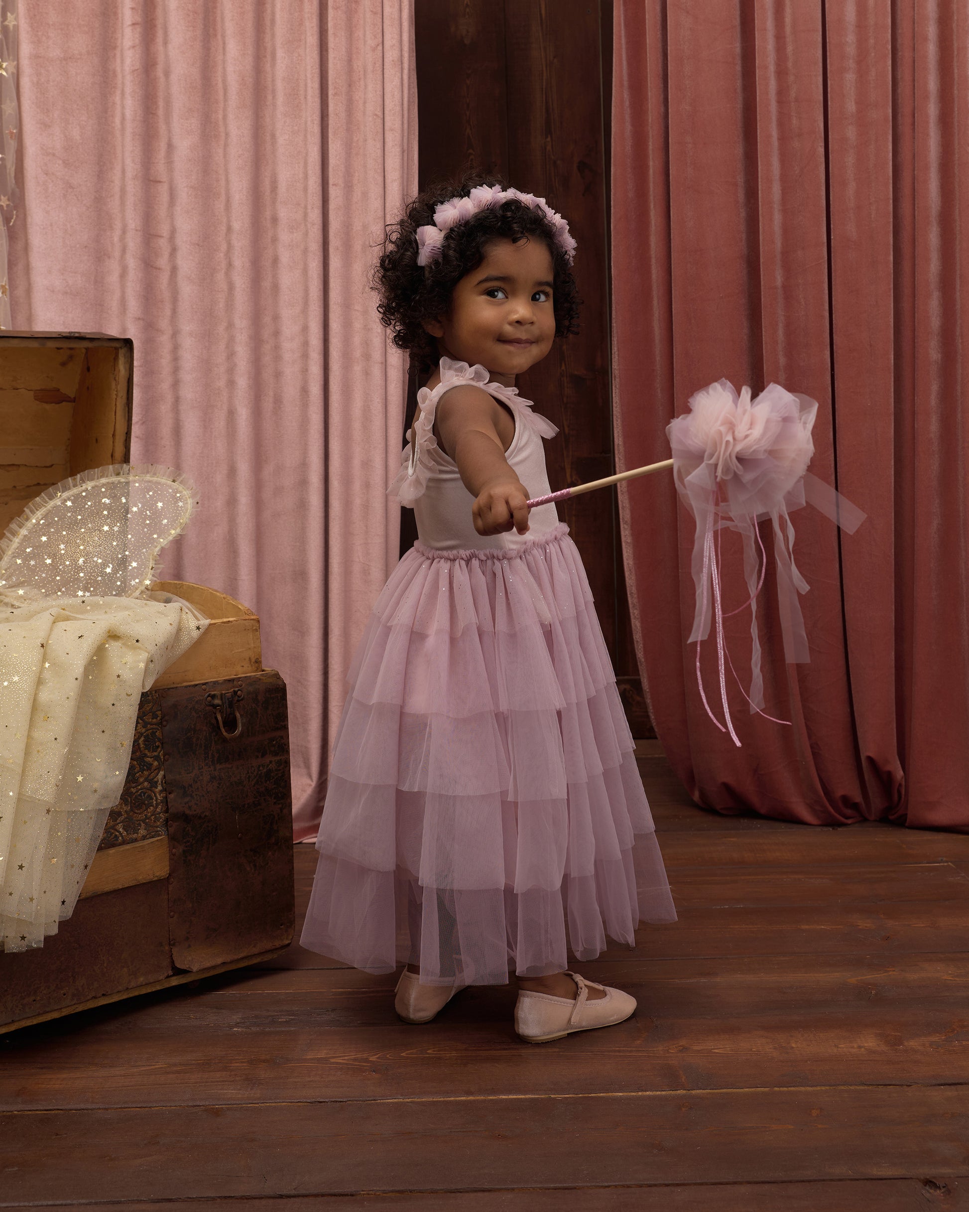 A young girl in a pink dress and headband holds the Noralee Pom Pom Crown + Wand, standing on a wooden floor with pink curtains and an open trunk in the background, ready for a magical dress-up adventure.