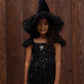 A young girl smiles at the camera in front of a wooden wall, wearing Noralee’s Witch Hat—a black, sparkly velvet accessory that adds a touch of magic to her dress-up adventure.
