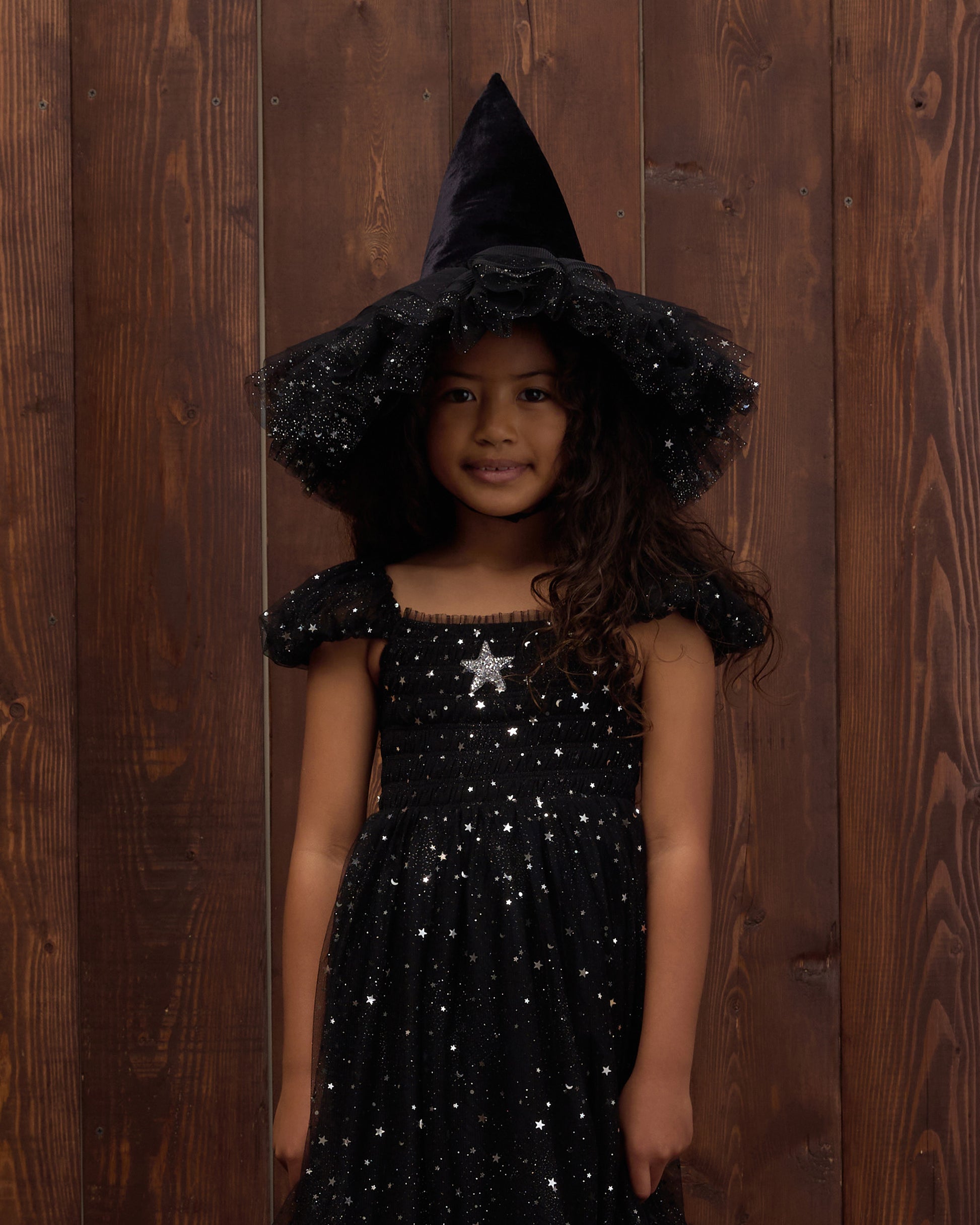 A young girl smiles at the camera in front of a wooden wall, wearing Noralee’s Witch Hat—a black, sparkly velvet accessory that adds a touch of magic to her dress-up adventure.
