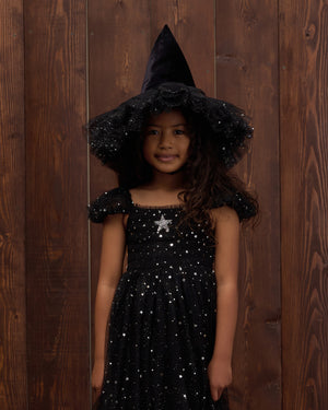 A young girl smiles at the camera in front of a wooden wall, wearing Noralee’s Witch Hat—a black, sparkly velvet accessory that adds a touch of magic to her dress-up adventure.