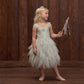 A young girl in Noralee’s Girls’ Magique Dress, a whimsical light blue sequin-embellished design, holds a wand and gazes to the side in front of a wooden wall.