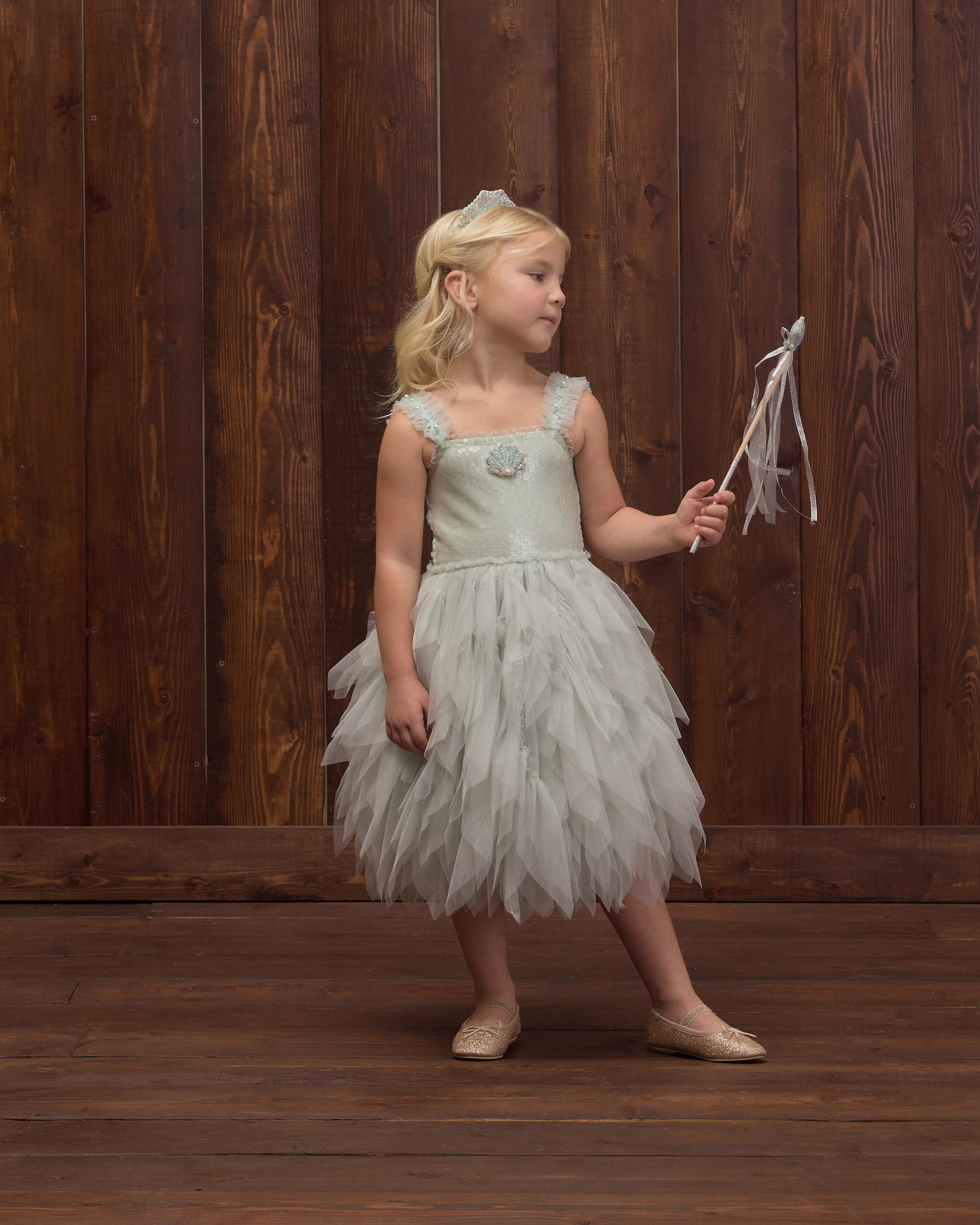 A young girl in Noralee’s Girls’ Magique Dress, a whimsical light blue sequin-embellished design, holds a wand and gazes to the side in front of a wooden wall.