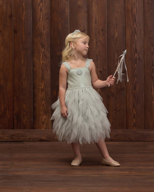 A young girl in Noralee’s Girls’ Magique Dress, a whimsical light blue sequin-embellished design, holds a wand and gazes to the side in front of a wooden wall.