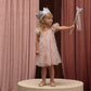 A young girl stands on a round platform wearing the Noralee Girls’ Edie Dress, featuring tulle sleeves and a gathered neckline. She is paired with a silver crown and holds a star wand, framed by pink and brown curtains in the background.