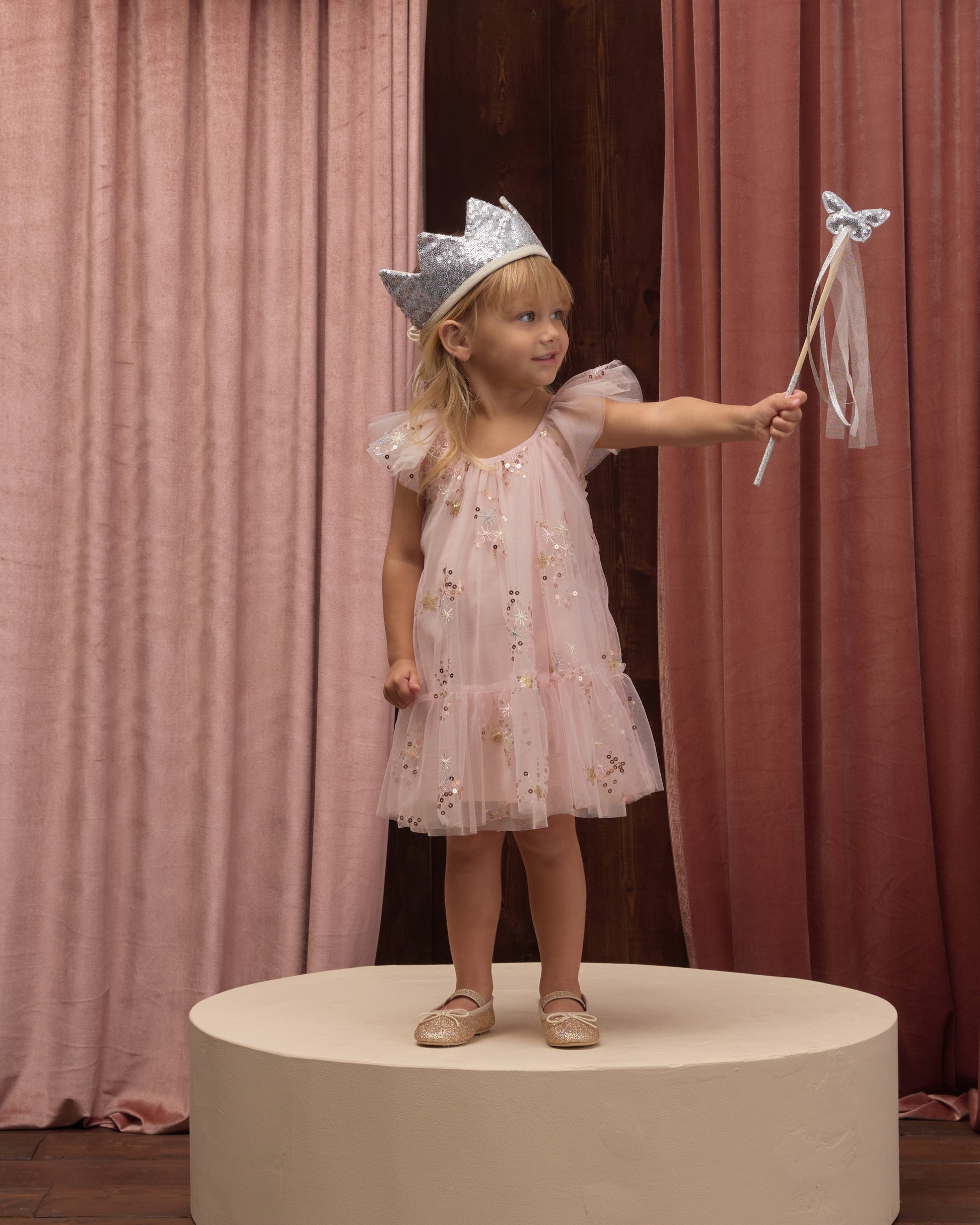 A young girl stands on a round platform wearing the Noralee Girls’ Edie Dress, featuring tulle sleeves and a gathered neckline. She is paired with a silver crown and holds a star wand, framed by pink and brown curtains in the background.