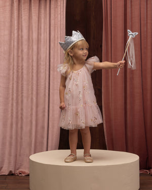 A young girl stands on a round platform wearing the Noralee Girls’ Edie Dress, featuring tulle sleeves and a gathered neckline. She is paired with a silver crown and holds a star wand, framed by pink and brown curtains in the background.