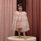 A young child wearing Noralee’s Tulle Cape is dressed in a layered pink dress with floral details, standing on a round platform before pink curtains, complemented by light shoes and a hair clip—an enchanting look perfect for dress-up or any special occasion.