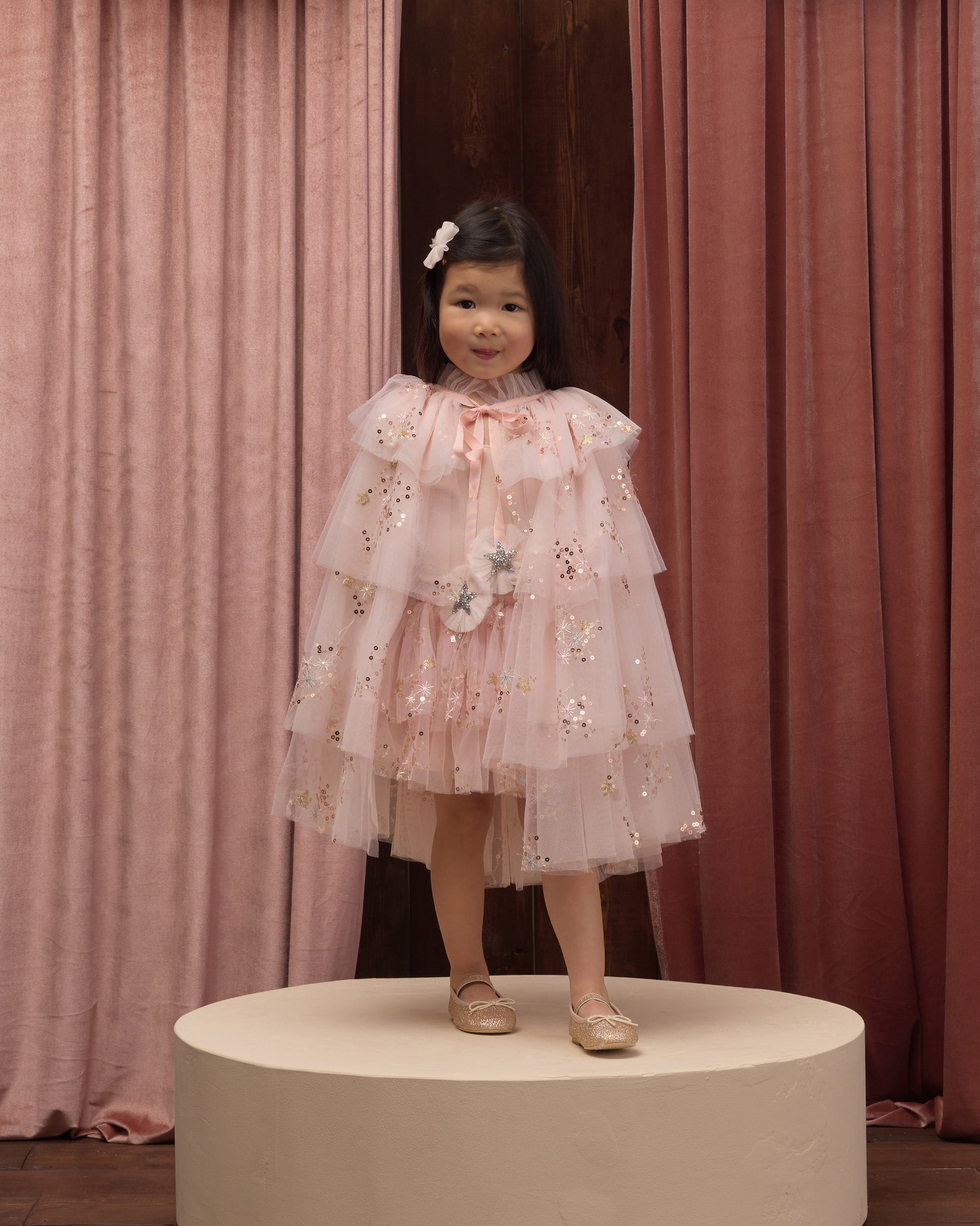 A young child wearing Noralee’s Tulle Cape is dressed in a layered pink dress with floral details, standing on a round platform before pink curtains, complemented by light shoes and a hair clip—an enchanting look perfect for dress-up or any special occasion.