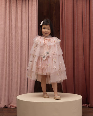 A young child wearing Noralee’s Tulle Cape is dressed in a layered pink dress with floral details, standing on a round platform before pink curtains, complemented by light shoes and a hair clip—an enchanting look perfect for dress-up or any special occasion.