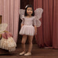 A young girl wears the Noralee Baby Girls’ and Girls’ Thea Tutu, standing on a wooden floor with velvet curtains and a box of dresses in the background.