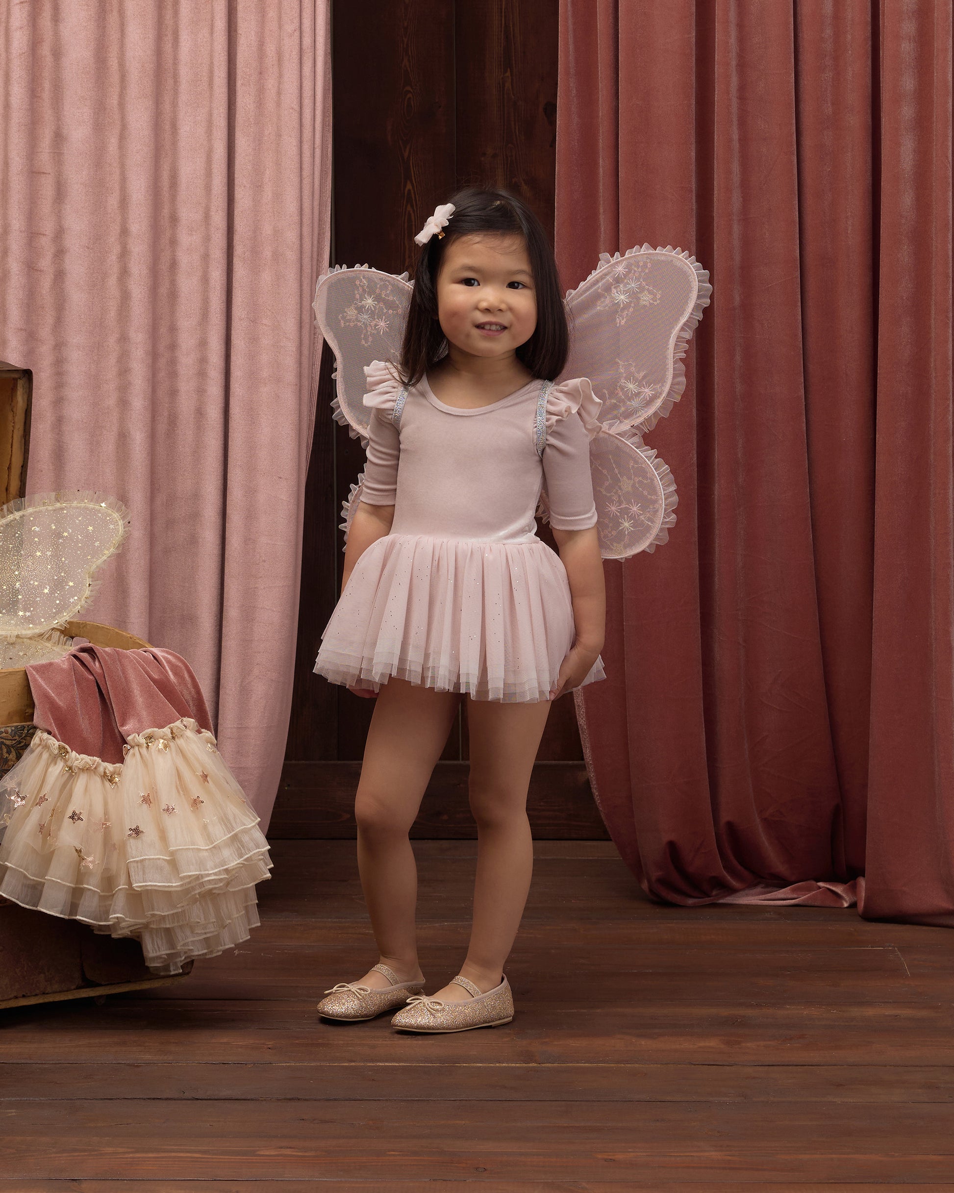 A young girl wears the Noralee Baby Girls’ and Girls’ Thea Tutu, standing on a wooden floor with velvet curtains and a box of dresses in the background.