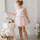 A young girl in a velvety soft leotard, pink tutu, and butterfly wings stands by a table with flowers, holding a wand and admiring a card. She dreams of joining the ranks of ballerinas like those wearing the Noralee Tullulah Hearts Tutu.