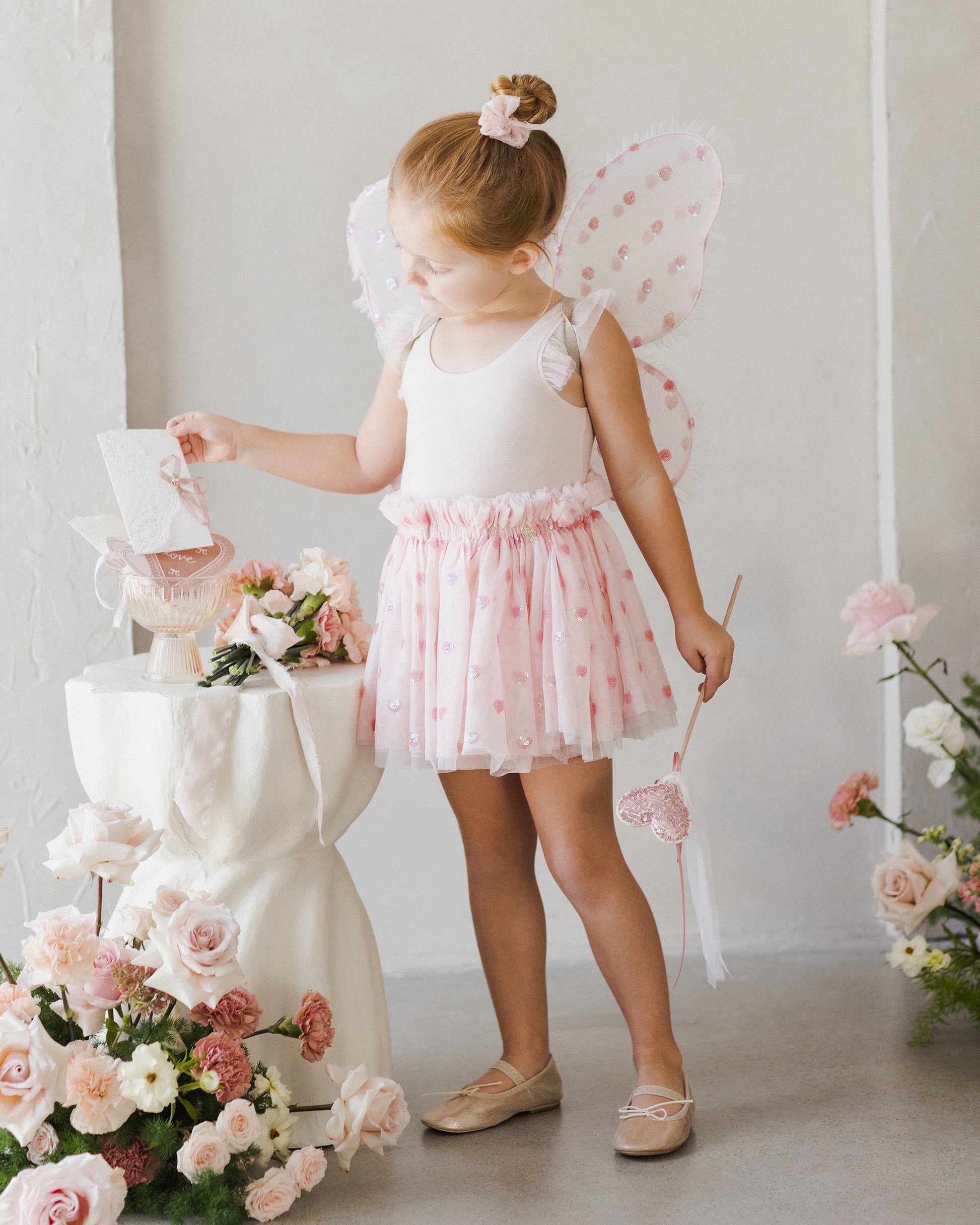 A young girl in a velvety soft leotard, pink tutu, and butterfly wings stands by a table with flowers, holding a wand and admiring a card. She dreams of joining the ranks of ballerinas like those wearing the Noralee Tullulah Hearts Tutu.