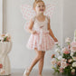 A young girl in the Noralee Blush Hearts Fairy Set, featuring a pink tutu and fairy wings, smiles indoors surrounded by pink flowers.