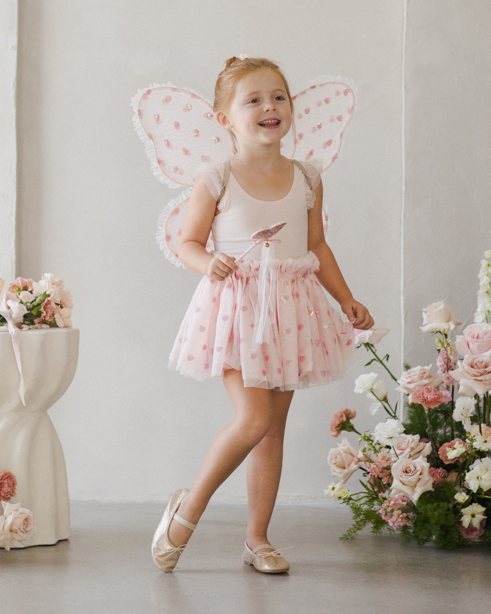 A young girl in the Noralee Blush Hearts Fairy Set, featuring a pink tutu and fairy wings, smiles indoors surrounded by pink flowers.