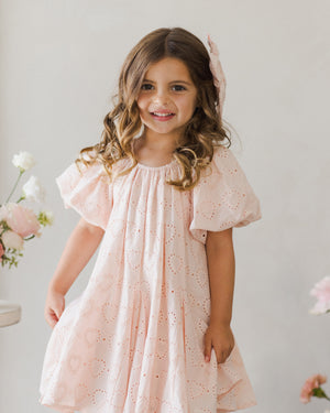 A young girl with long hair beams next to light-colored flowers, wearing a blush-hued Noralee Maia Dress decorated with charming heart patterns.