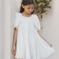 A young girl wearing the Noralee Baby Girls’ and Girls’ Maia Dress, a white embroidered heart eyelet mini dress, stands by a light-colored wall, gently holding the side of her dress while looking down. Shrubs can be seen in the background.
