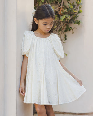 A young girl wearing the Noralee Baby Girls’ and Girls’ Maia Dress, a white embroidered heart eyelet mini dress, stands by a light-colored wall, gently holding the side of her dress while looking down. Shrubs can be seen in the background.