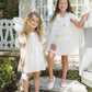 Two young girls stand outdoors on stone steps, dressed in Noralee’s Baby Girls’ and Girls’ Lottie Tutu Set: white, fairy-inspired costumes featuring double-tiered tulle tutus and flutter sleeve leotards, with matching capes and wings catching the light in front of a white gate and sunlit garden.