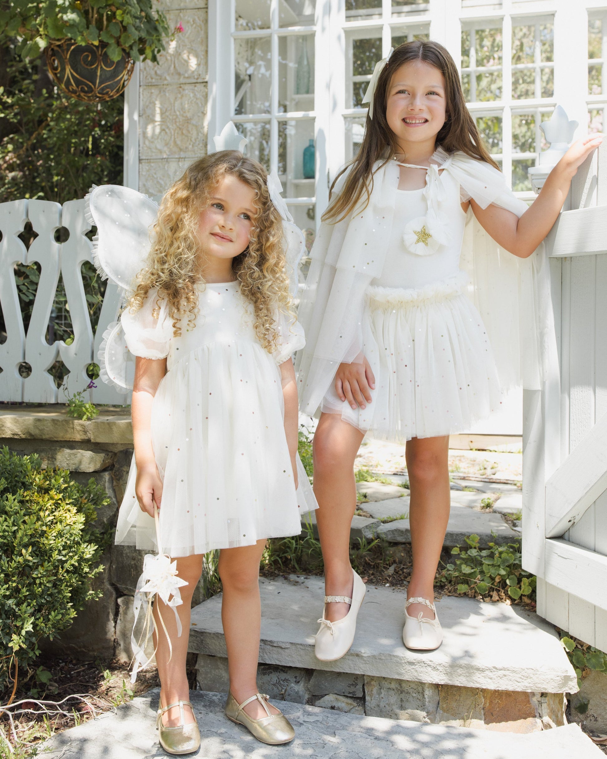 Two young girls stand outdoors on stone steps, dressed in Noralee’s Baby Girls’ and Girls’ Lottie Tutu Set: white, fairy-inspired costumes featuring double-tiered tulle tutus and flutter sleeve leotards, with matching capes and wings catching the light in front of a white gate and sunlit garden.