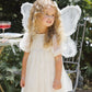 A young child with curly blonde hair wears a charming white dress with small dots and large white fairy wings from the Noralee Girls’ Fairy Set, standing outdoors near a metal chair and table for an enchanting dress-up moment.