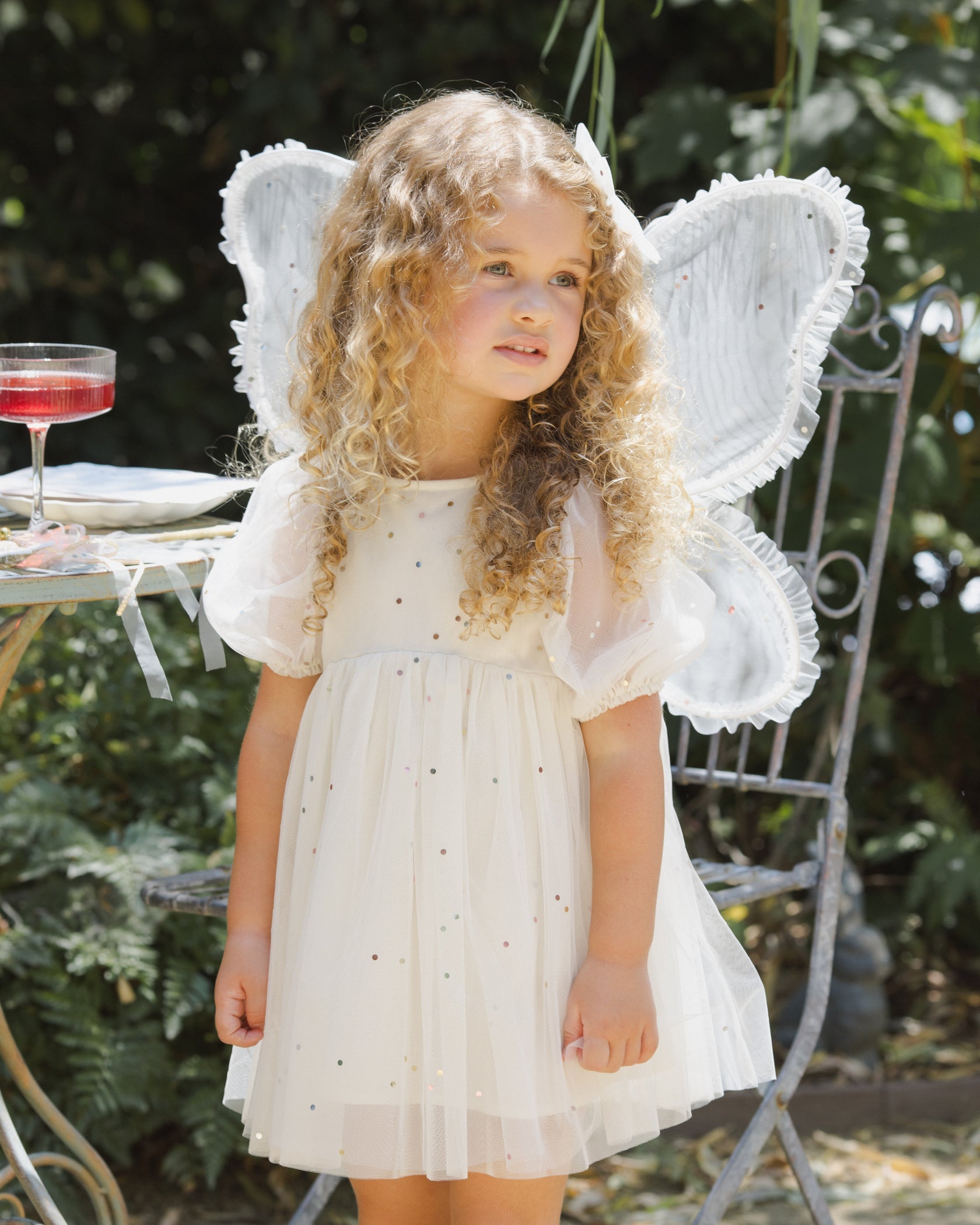 A young child with curly blonde hair wears a charming white dress with small dots and large white fairy wings from the Noralee Girls’ Fairy Set, standing outdoors near a metal chair and table for an enchanting dress-up moment.