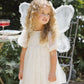 A young child with curly hair wears Noralee’s Girls’ Luna Dress—a cream babydoll mini dress featuring small dots and delicate puff sleeves—paired with white fairy wings, standing outdoors beside a metal chair and table.