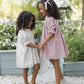 Two young girls wearing Noralee’s Girls’ Maddie Dress stand hand in hand on a gravel path, smiling at each other amidst a garden full of blooming flowers and lush greenery.