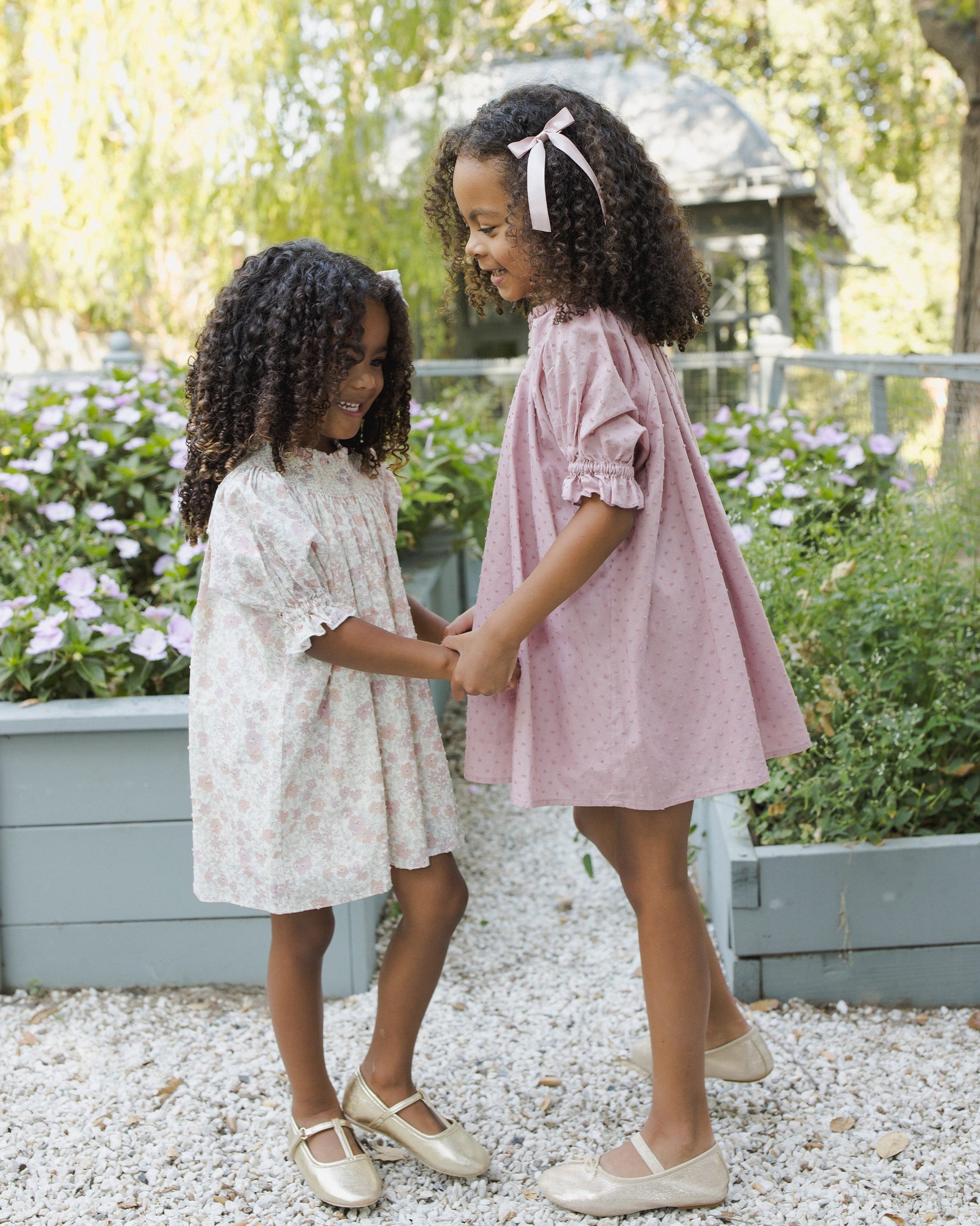 Two young girls wearing Noralee’s Girls’ Maddie Dress stand hand in hand on a gravel path, smiling at each other amidst a garden full of blooming flowers and lush greenery.
