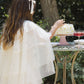 Wearing the Noralee Girls’ Tulle Cape over her white dress and a matching hair ribbon, a young girl places a gold glitter star atop a cake outdoors, with a pink drink beside her—capturing the magic of every party princess.