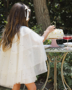 Wearing the Noralee Girls’ Tulle Cape over her white dress and a matching hair ribbon, a young girl places a gold glitter star atop a cake outdoors, with a pink drink beside her—capturing the magic of every party princess.