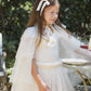 Outdoors, a young girl embodies the party princess look as she stands by a table set with drinks and plates, wearing the Noralee Girls’ Tulle Cape adorned with glittering stars and styled with a ribbon in her hair, surrounded by lush greenery.