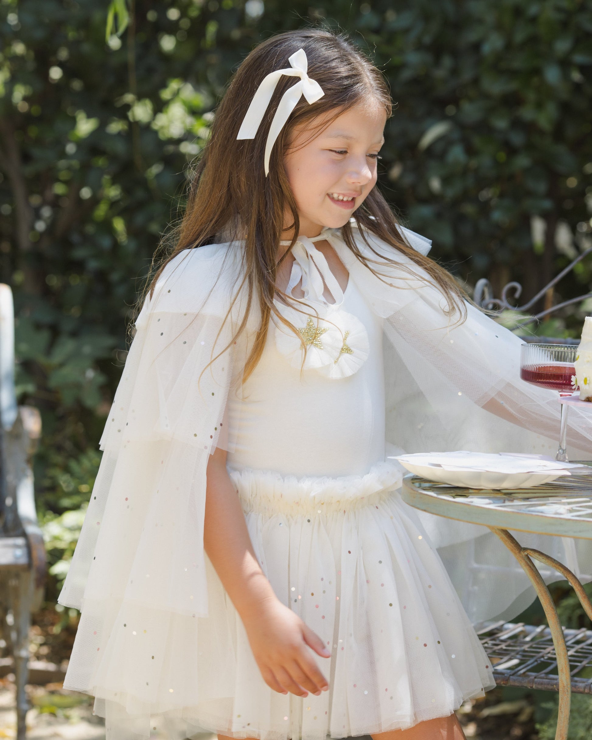 Outdoors, a young girl embodies the party princess look as she stands by a table set with drinks and plates, wearing the Noralee Girls’ Tulle Cape adorned with glittering stars and styled with a ribbon in her hair, surrounded by lush greenery.