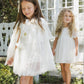 Two young girls in white dresses stand outdoors near a white gate and glass doors—one, dressed as a party princess, wears the Noralee Girls’ Tulle Cape adorned with gold glitter stars and a matching star pin, while the other has curly hair and fairy wings.