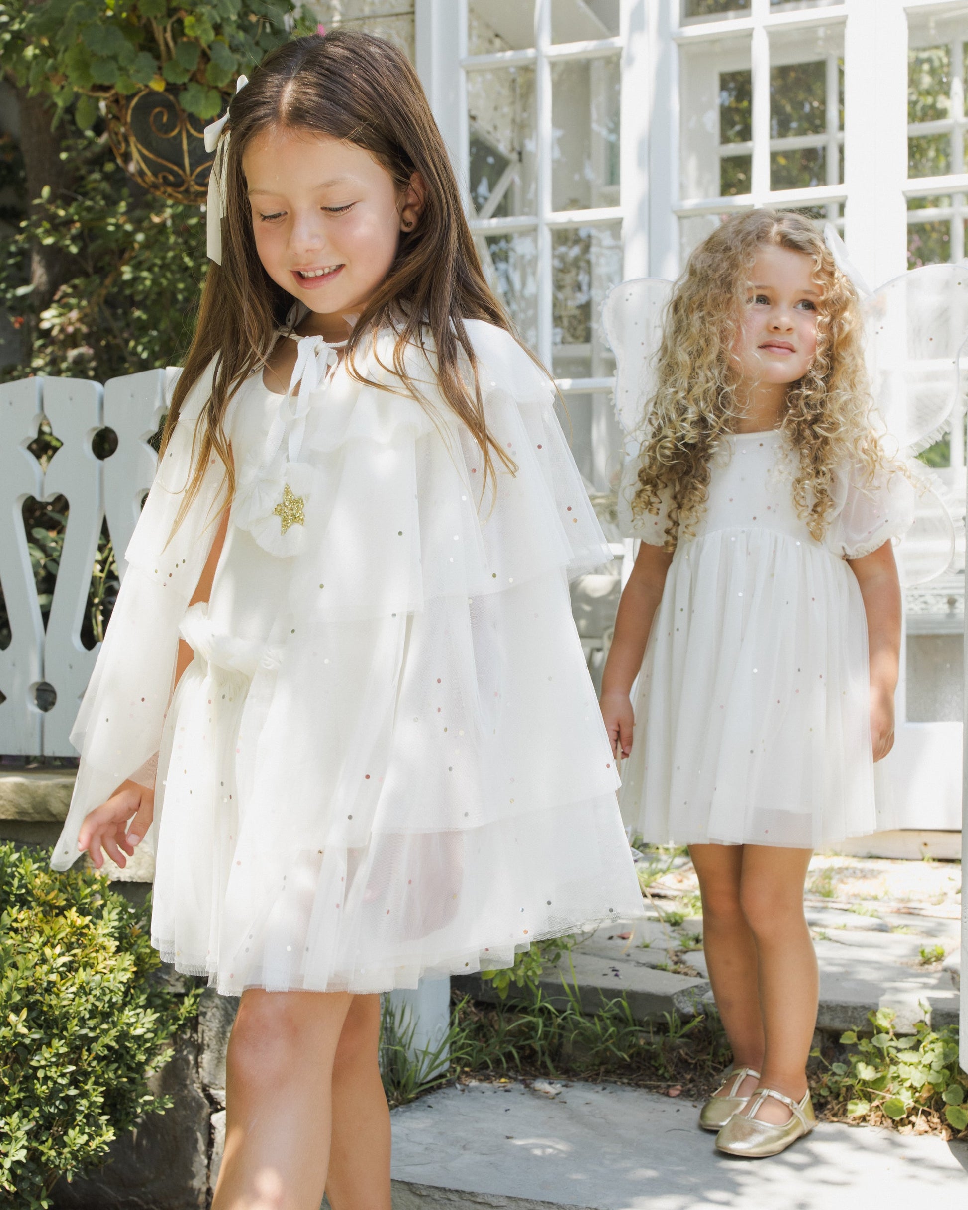 Two young girls in white dresses stand outdoors near a white gate and glass doors—one, dressed as a party princess, wears the Noralee Girls’ Tulle Cape adorned with gold glitter stars and a matching star pin, while the other has curly hair and fairy wings.