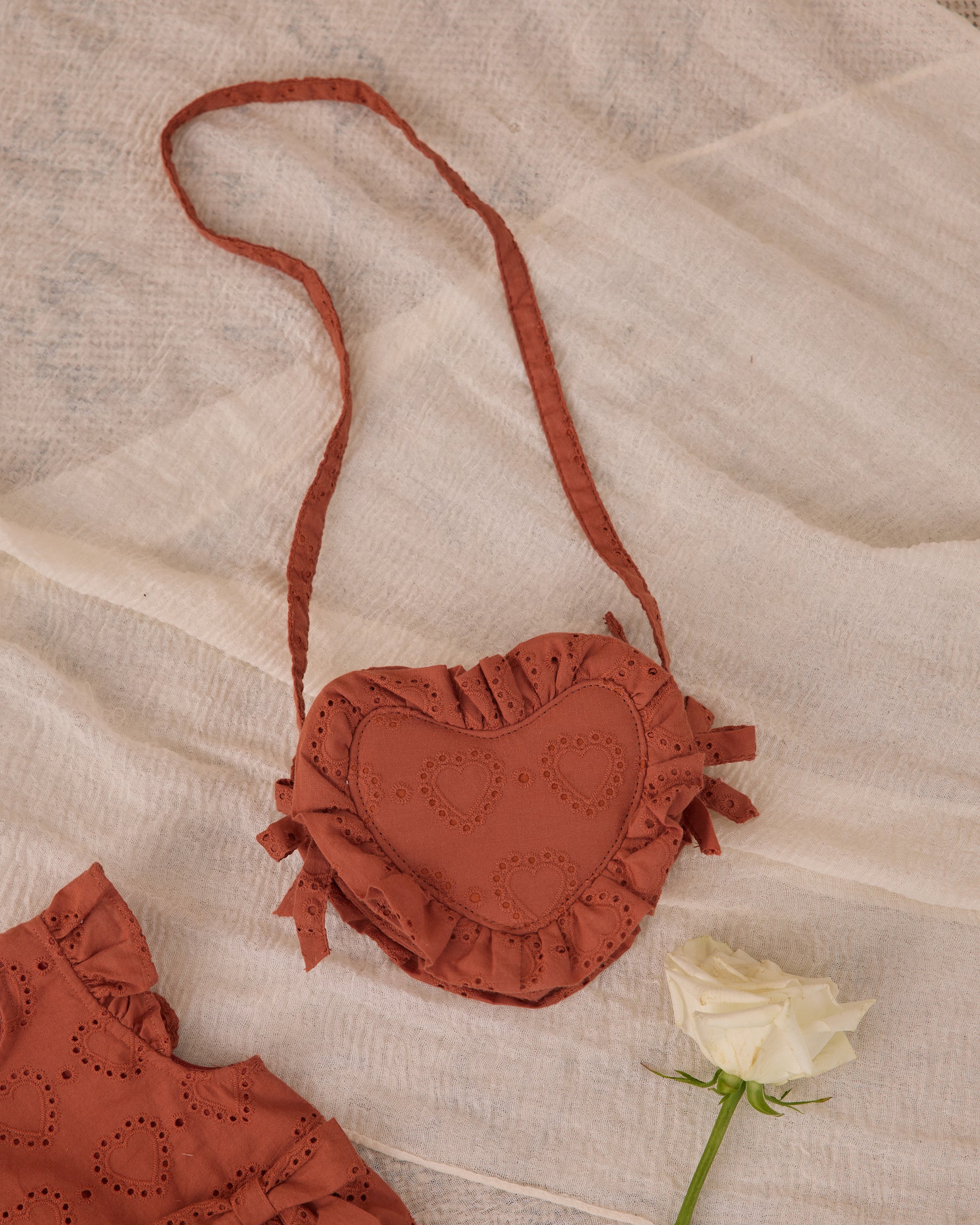 The Noralee Girls' Heart Purse, crafted from rust heart eyelet fabric with ruffled edges, is displayed on a cream cloth beside a white rose—a charming accessory for girls.