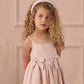 A young girl with curly blonde hair and a light pink headband wears the Noralee Girls' Beatrice Midi Dress, featuring smocking and bows, standing before a beige paneled wall.