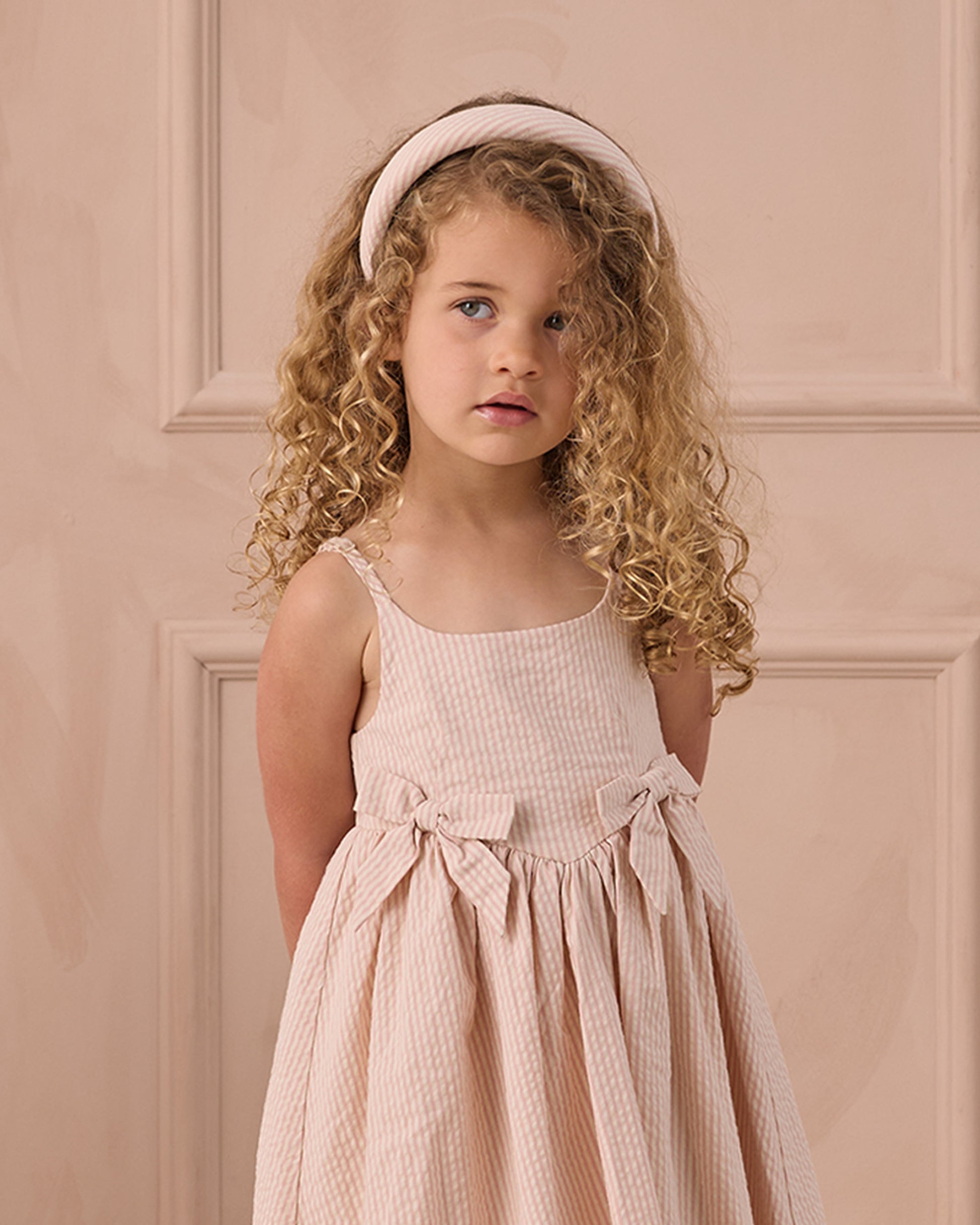 A young girl with curly blonde hair and a light pink headband wears the Noralee Girls' Beatrice Midi Dress, featuring smocking and bows, standing before a beige paneled wall.