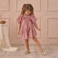 A young girl stands indoors on a patterned rug wearing the Noralee Girls' Maia Dress—pink with heart eyelet details and puff sleeves—paired with gold shoes, looking down as she extends one leg.