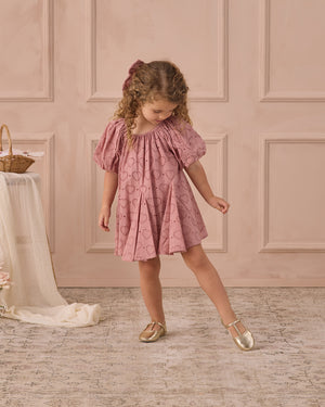 A young girl stands indoors on a patterned rug wearing the Noralee Girls' Maia Dress—pink with heart eyelet details and puff sleeves—paired with gold shoes, looking down as she extends one leg.